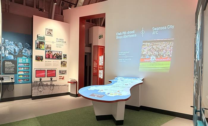 View into a white gallery with red trim, video projection onto a wall and table, photographs and text on the walls, glass display cases and a green tu